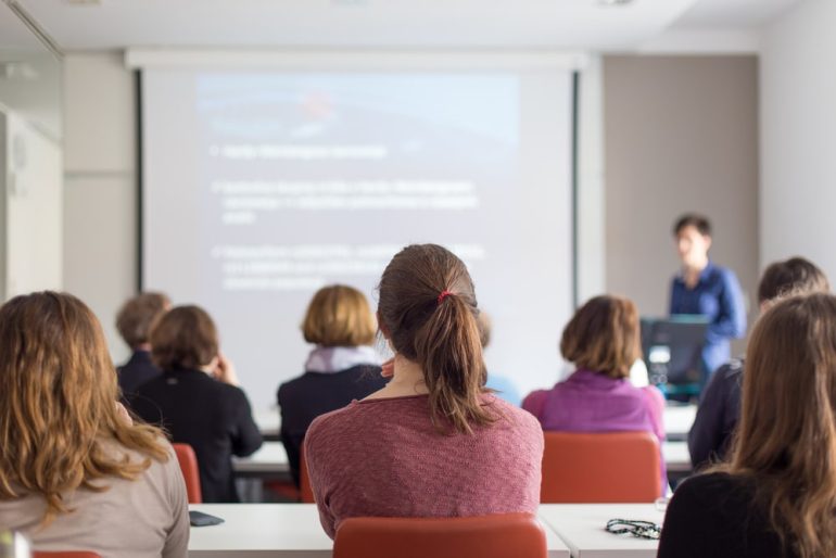 Female speaker giving presentation in lecture hall at university workshop. Rear view of unrecognized participants listening to lecture and making notes. Scientific conference event.