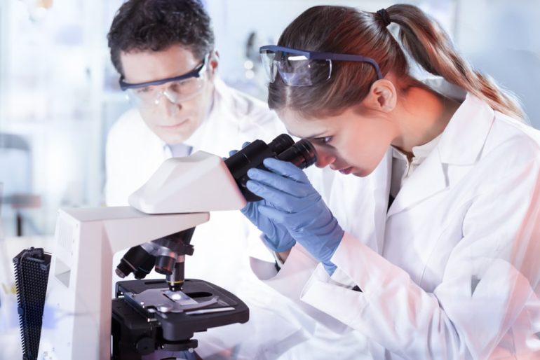 Life scientists researching in laboratory. Attractive female young scientist and her doctoral supervisor microscoping in their working environment. Healthcare and biotechnology.